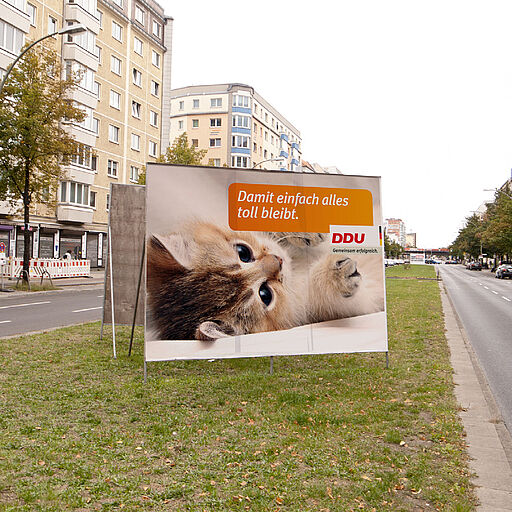 Deutsch – Bildbeschreibung (SEO / Metadaten) Plakatmotiv aus einer experimentellen Plakatintervention im öffentlichen Raum auf der Frankfurter Allee. Zu sehen ist ein Motiv mit einem Babykätzchen, darüber der Schriftzug „Damit einfach alles toll bleibt“ sowie der Parteiname „DDU – gemeinsam erfolgreich“, als bewusste Anlehnung an bekannte Wahlwerbung. Das Plakat wurde ungefragt zwischen reguläre Werbeplakate geklebt und nutzt vertraute politische Bildsprache, um deren Mechanismen im Stadtraum sichtbar zu machen. ⸻ English – Image description (SEO / metadata) Poster from an experimental public-space poster intervention on Frankfurter Allee in Berlin. The visual features a cute kitten, accompanied by the slogan “So that everything simply stays great” and the fictional party name “DDU – together successful,” deliberately referencing established election campaign aesthetics. The poster was placed without authorization among regular outdoor advertising, examining the visual language of political communication in public space.