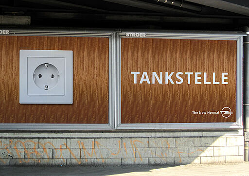 Deutsch – Bildbeschreibung (SEO / Google-Kampagne) Plakatmotiv der Google-Kampagne „The New Normal“ für Opel. Auf einem schlichten Holzhintergrund ist links eine normale Haushalts-Steckdose zu sehen, rechts der Schriftzug „Tankstelle“. Reduziertes Motiv mit humorvollem Perspektivwechsel zur Elektromobilität im Alltag. ⸻ English – Image description (SEO / Google campaign) Poster visual from the Google campaign “The New Normal” for Opel. Set against a plain wooden background, a standard household power socket is shown on the left, with the word “Tankstelle” on the right. Minimal visual using humor to reframe everyday electric mobility.